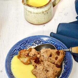 A bowl of chocolate chip apple crumble with custard and jug of custard behind.