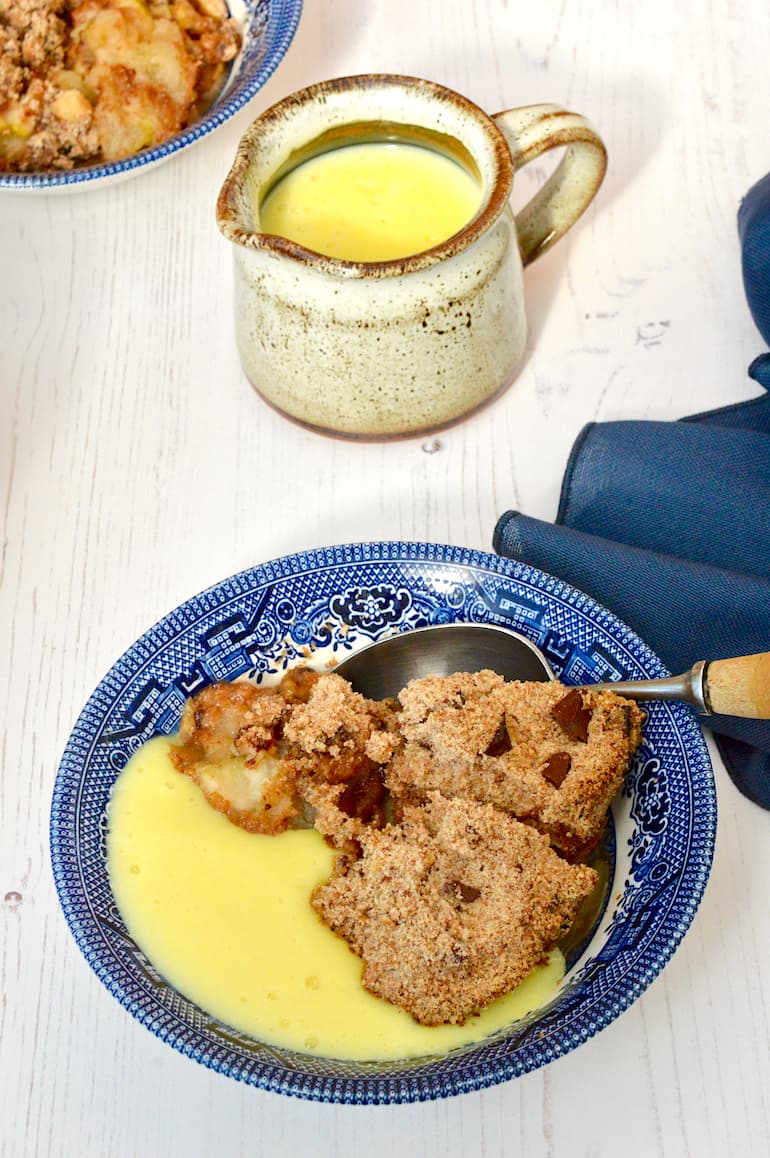 A bowl of chocolate chip apple crumble with custard and jug of custard behind.