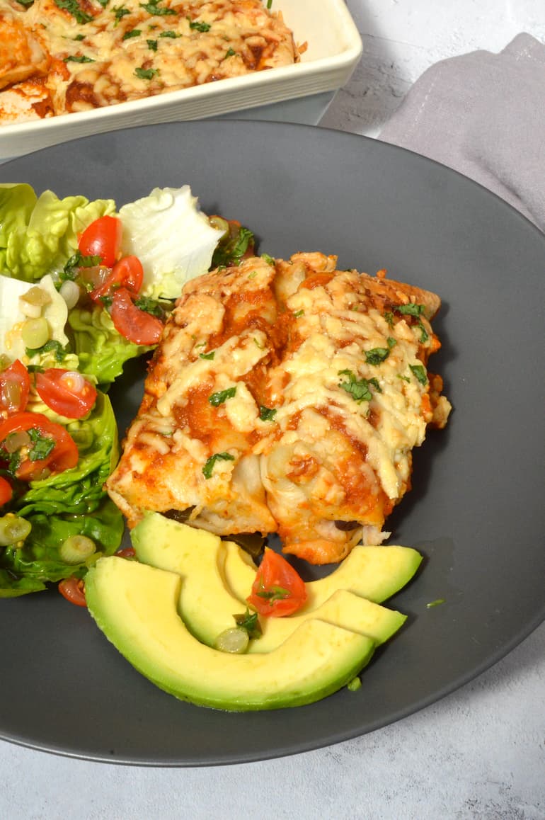 Two vegetarian enchiladas on a grey plate with avocado and salad.