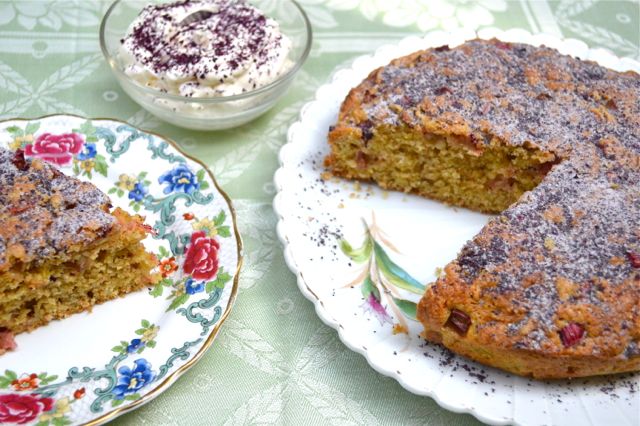 Remarkable Rose Rhubarb Cake sitting on a plate with one slice removed to a side plate and a bowl of rose cream in the background.