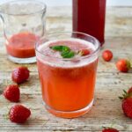 Glass of homemade strawberry rose mint fizz with strawberries and bottle of rose syrup.
