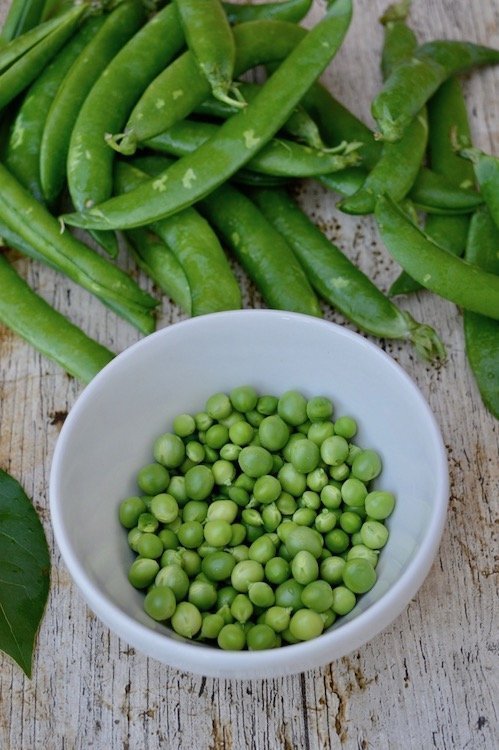 Podded Peas in a bowl with pods and a bay leaf in the background.