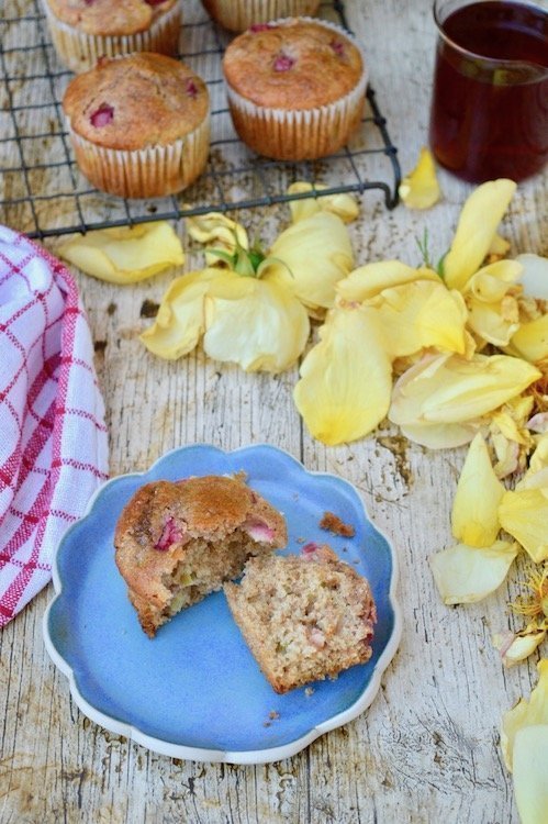 A halved rhubarb muffin on a blue plate with rose petals, rose syrup and spelt rhubarb muffins in the background