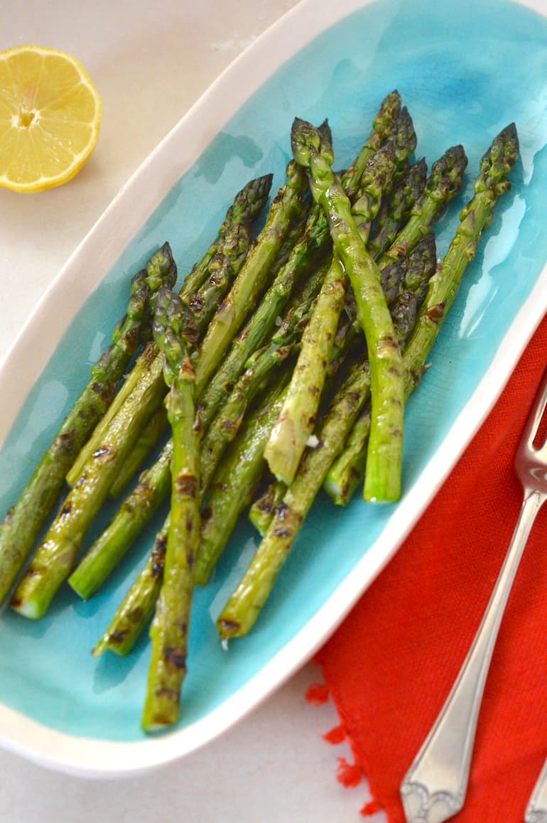 Turquoise serving plate with griddled asparagus, lemon halves, napkins and forks on the sides.