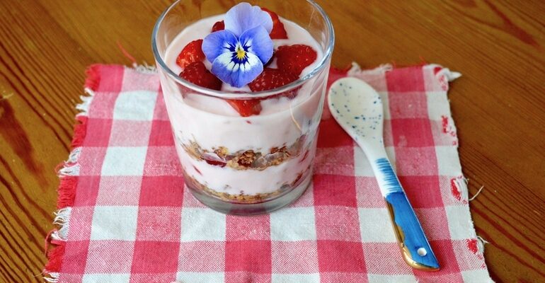Raspberry granola parfait in a glass with a blue pansy on top and a spoon by the side.