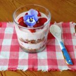 Raspberry granola parfait in a glass with a blue pansy on top and a spoon by the side.
