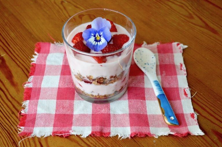 Raspberry granola parfait in a glass with a blue pansy on top and a spoon by the side.