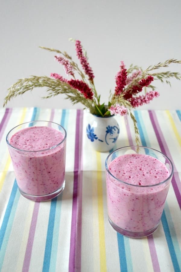 Two glasses of blueberry kefir smoothie with a vase of flowers in the background.