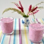 Two glasses of blueberry kefir smoothie with a vase of flowers in the background.