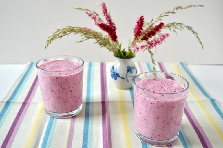 Two glasses of blueberry kefir smoothie with a vase of flowers in the background.