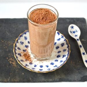 A glass of salted caramel chocolate milkshake sitting on a blue and white plate with a blue and white spoon alongside.