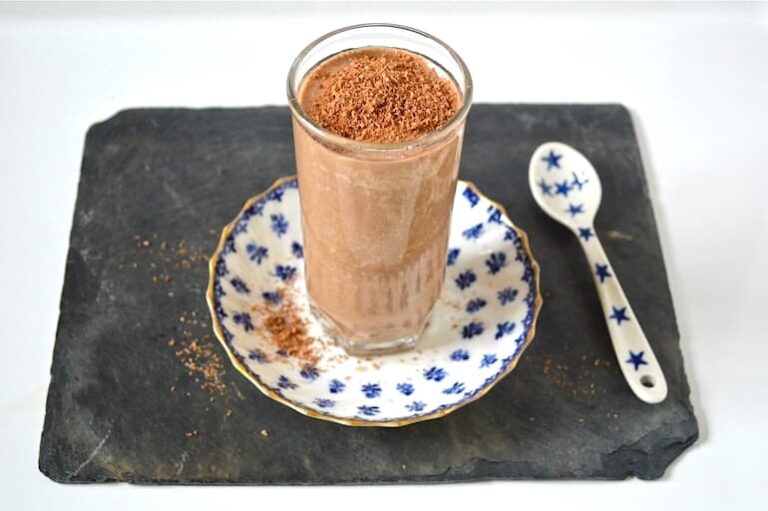 A glass of salted caramel chocolate milkshake sitting on a blue and white plate with a blue and white spoon alongside.