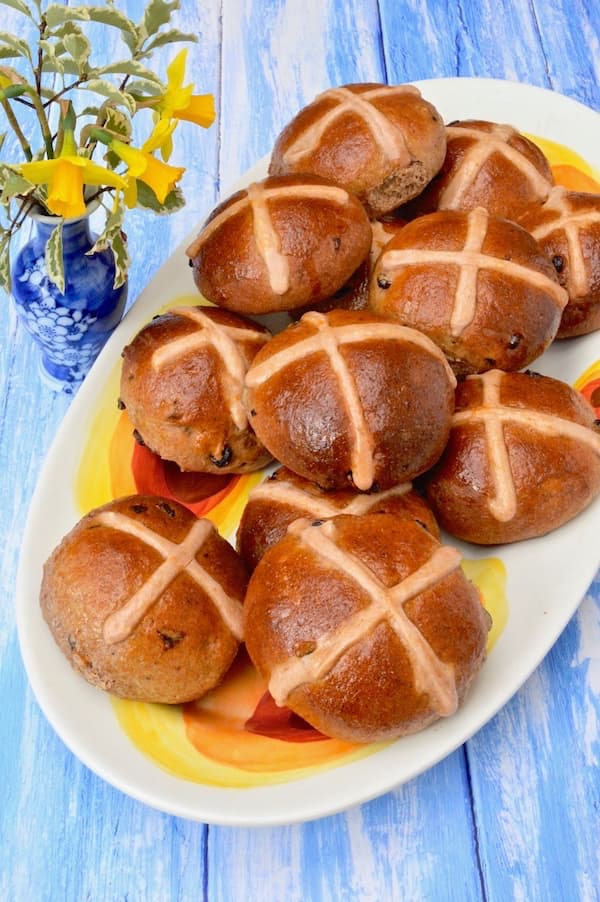 Vegan hot cross buns piled on a platter with a little vase of daffodils on the side.