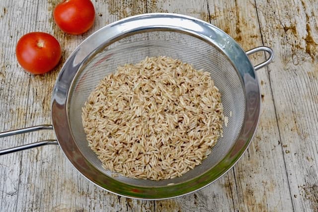 Long grain brown rice draining in a sieve with two tomatoes in the background.