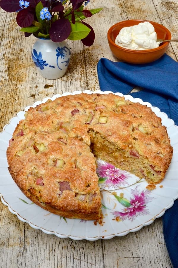 Rhubarb and ginger cake on a plate with one slice removed. A bowl of whipped cream, a vase of flowers and a blue napkin are in the background.