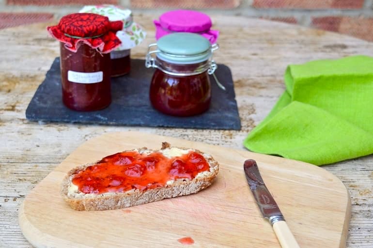 A slice of bread spread with butter and easy homemade strawberry jam, with jars of it in the background.