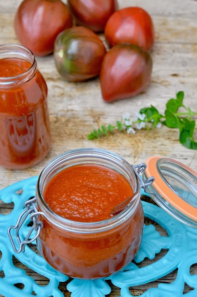 An open jar of homemade Easy Tomato Sauce with another open jar in the background along with whole fresh tomatoes and a sprig of flowering basil.