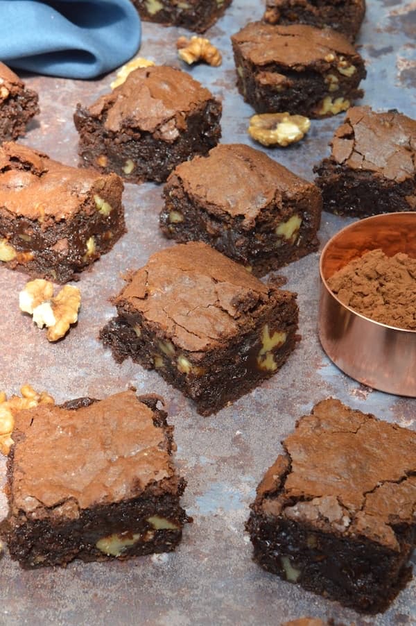 Real chocolate brownies on a baking tray with walnuts scattered about, a pot of cocoa powder and a blue napkin in the background.