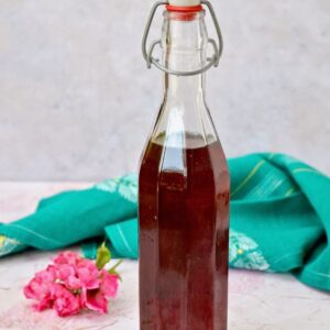 Bottle of homemade rose syrup with a turquoise cloth and pink rose in the background.