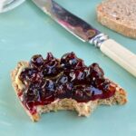 Easy to make blackcurrant chilli jam spread on a slice of bread with a butter knife and jar of blackcurrant jam in the background.