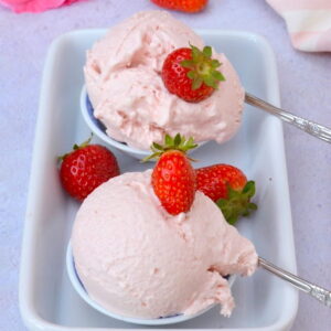 Two mini bowls each holding a scoop of easy no-churn strawberry ice cream. Includes fresh strawberries, a rose and two apostle spoons.