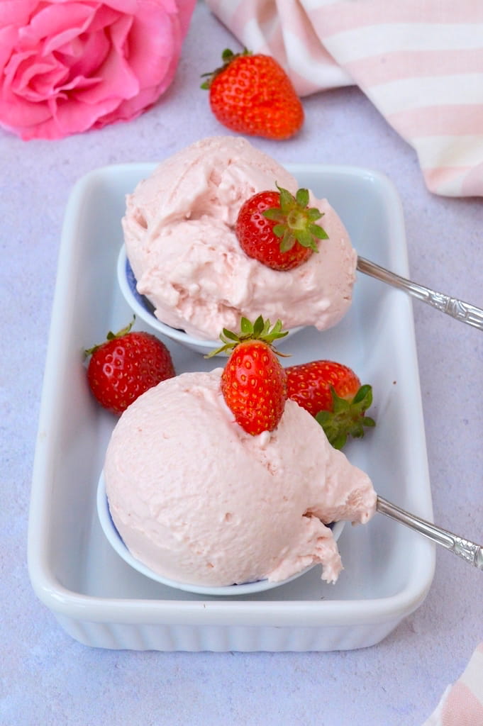 Two mini bowls each holding a scoop of easy no-churn strawberry ice cream. Includes fresh strawberries, a rose and two apostle spoons.