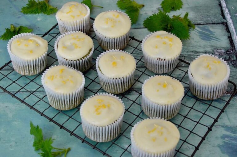 A rack of lemon and white chocolate cupcakes cooling on a wire rack with a few green nettle tops scattered about.