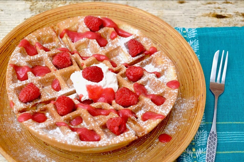 Wholemeal spelt waffles with rose raspberry sauce on a bamboo platter with a fork and blue napkin to the side.