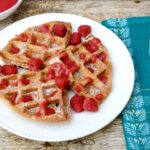 Wholemeal spelt waffles with rose raspberry sauce on white plate with fork and blue napkin to side.
