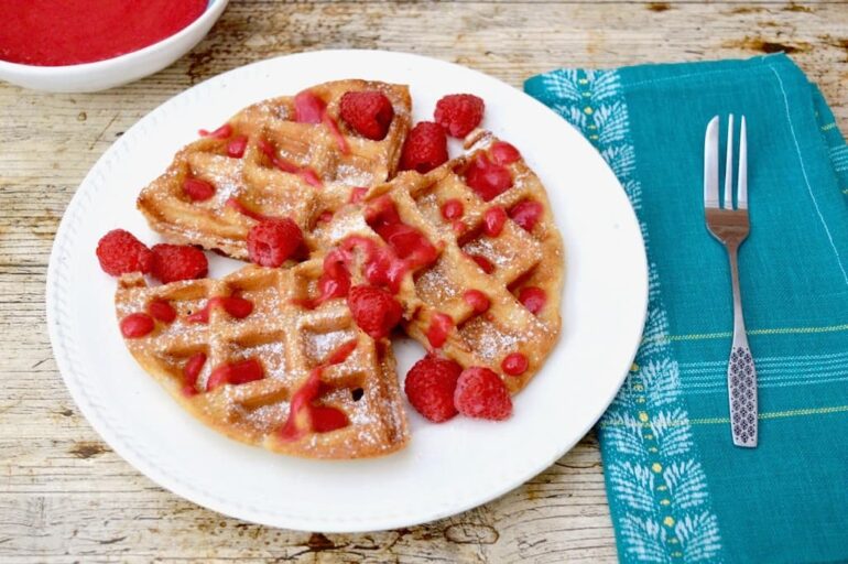 Wholemeal spelt waffles with rose raspberry sauce on a white plate with a fork and blue napkin to the side.