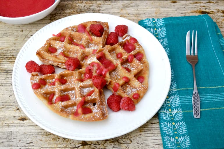 Wholemeal spelt waffles with rose raspberry sauce on white plate with fork and blue napkin to side.