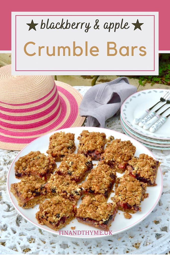 Autumn fruit crumble bars on a cake stand in the garden with a striped pink straw hat in the background. Text box reads "blackberry & apple crumble bars".