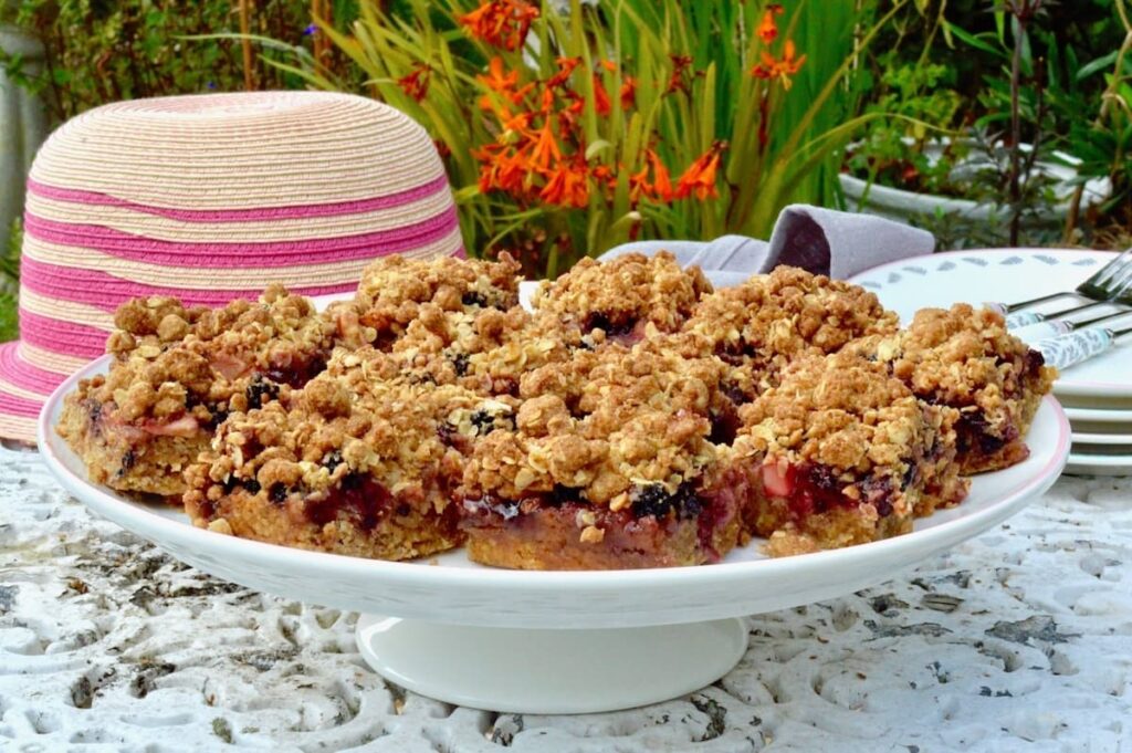Blackberry and apple crumble bars on a cake stand in the garden with a striped pink straw hat in the background.