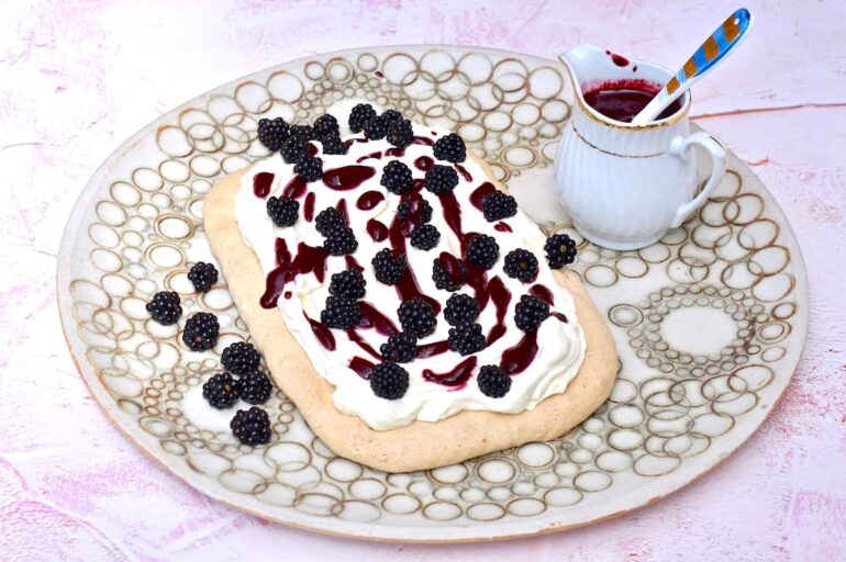 Rectangular hazelnut meringue with blackberries on a round pottery platter along with a jug of blackberry sauce.