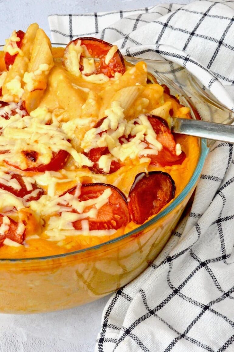 A dish of healthy pumpkin mac and cheese topped with tomatoes. Serving spoon lifting from the back. and served with cabbage.