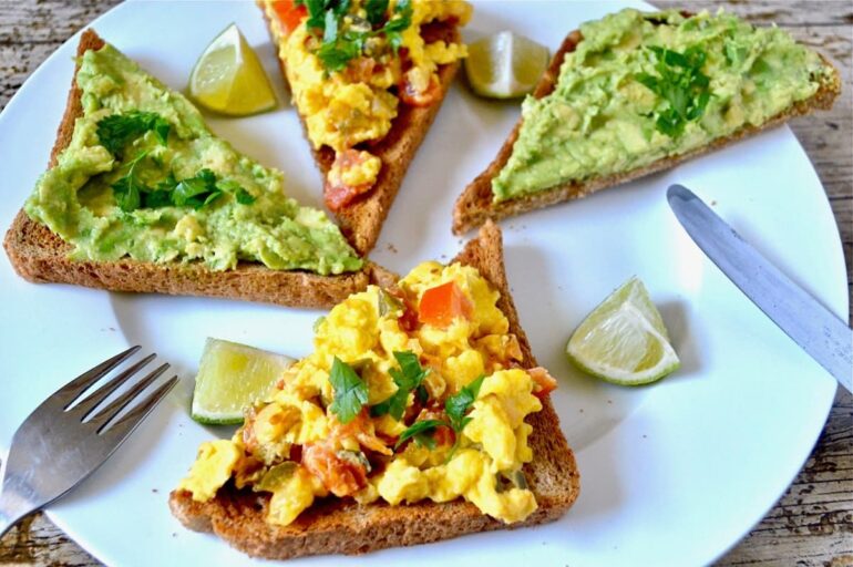 Slices of toast topped with Huevos Rancheros or Smashed Avocado. On a white plate with lime segments and a knife and fork.