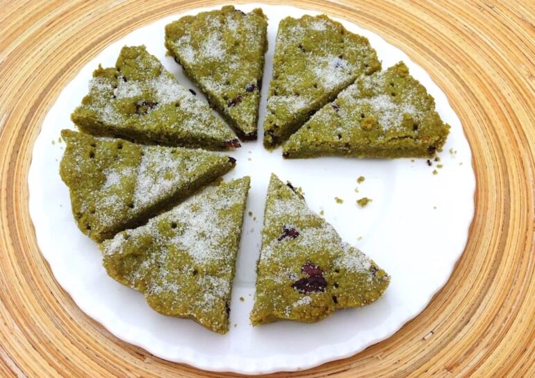 Triangles of cacao nibbed matcha shortbread on a white plate with one piece missing.