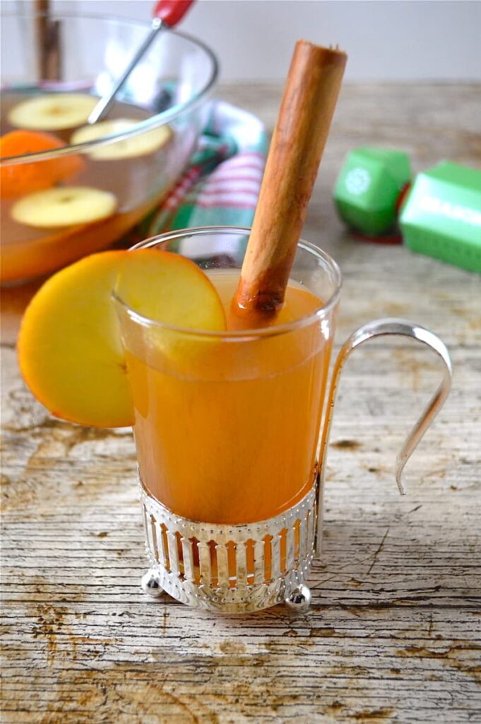 A glass of warm mulled cider or apple juice with a slice of apple on the side and a cinnamon stick to stir.