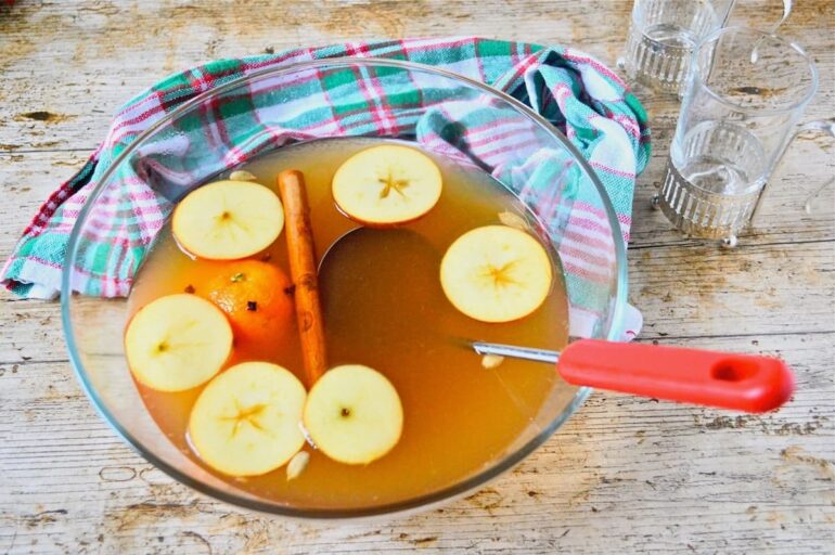 A glass bowl of mulled cider or mulled apple juice with floating apple slices, clove studded orange and cinnamon stick.