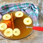 Glass bowl of mulled cider with floating apple slices, clove studded orange and cinnamon stick.