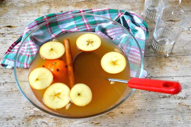 Glass bowl of mulled cider with floating apple slices, clove studded orange and cinnamon stick.