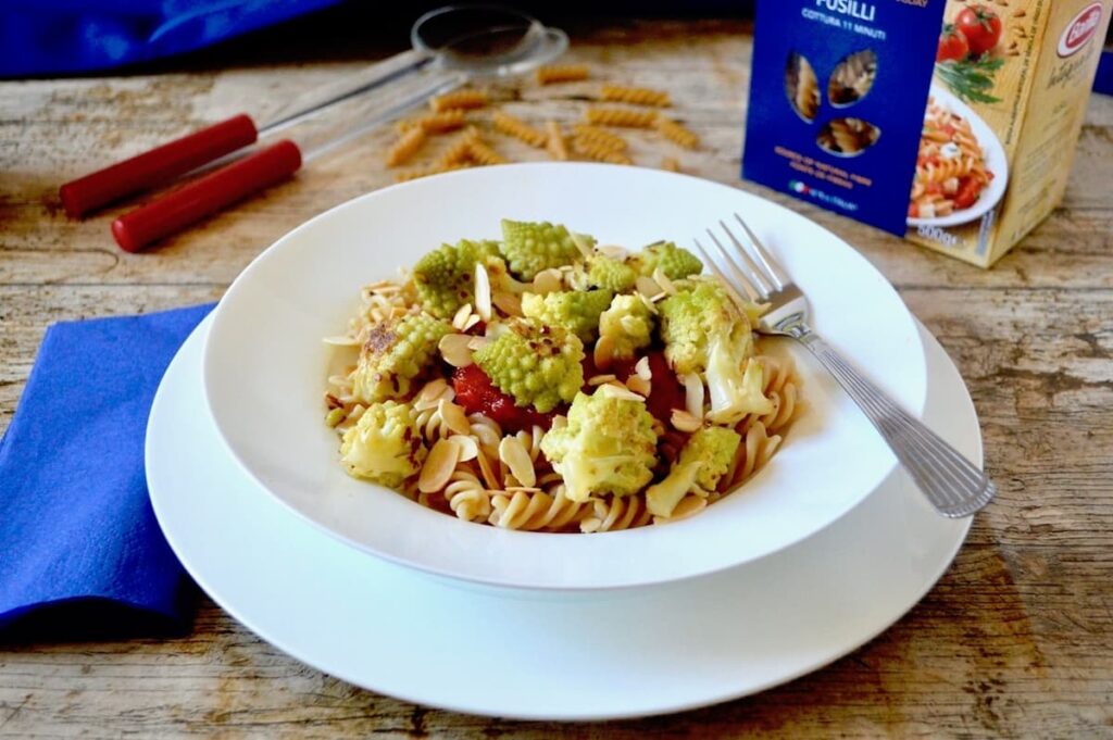 A bowl of romanesco pasta with fork, sitting on a plate. Blue napkin, servers and a box of pasta in the background.