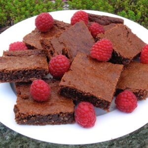 A pile of gluten-free almond toffee brownies with raspberries scattered over them sitting on a white plate.