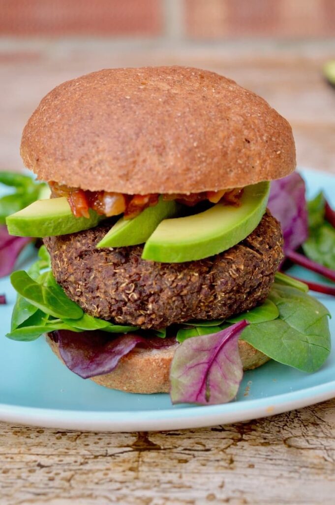 Red quinoa chickpea burger in a wholemeal bun with avocado slices, tomato and onion relish and salad leaves.