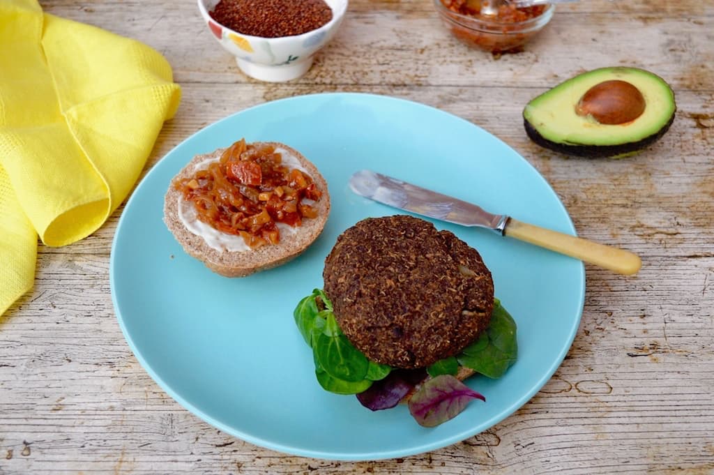 Red quinoa burger with black chickpeas and prunes on a half bun with salad. Caramelised onion and tomato relish with mayonnaise on other bun half.