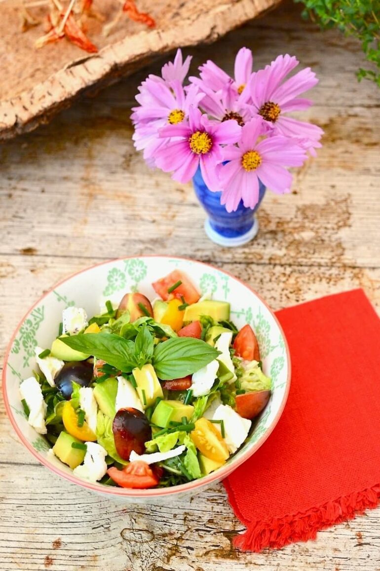 A bowl of caprese salad with avocado and lettuce. A vase of pink flowers in the background.