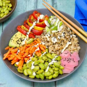 Vegan sushi bowl with brown rice, pickled ginger, edamame beans, carrots, avocado, peppers and onions. Chopsticks resting on the side and a bowl of edamame beans with blue napkin at the rear.