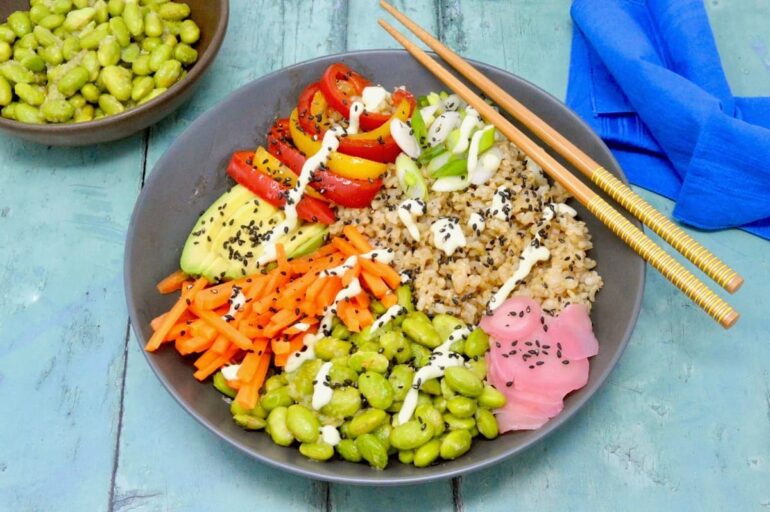 Vegan sushi bowl with brown rice, pickled ginger, edamame beans, carrots, avocado, peppers and onions. Chopsticks resting on the side and a bowl of edamame beans with blue napkin at the rear.
