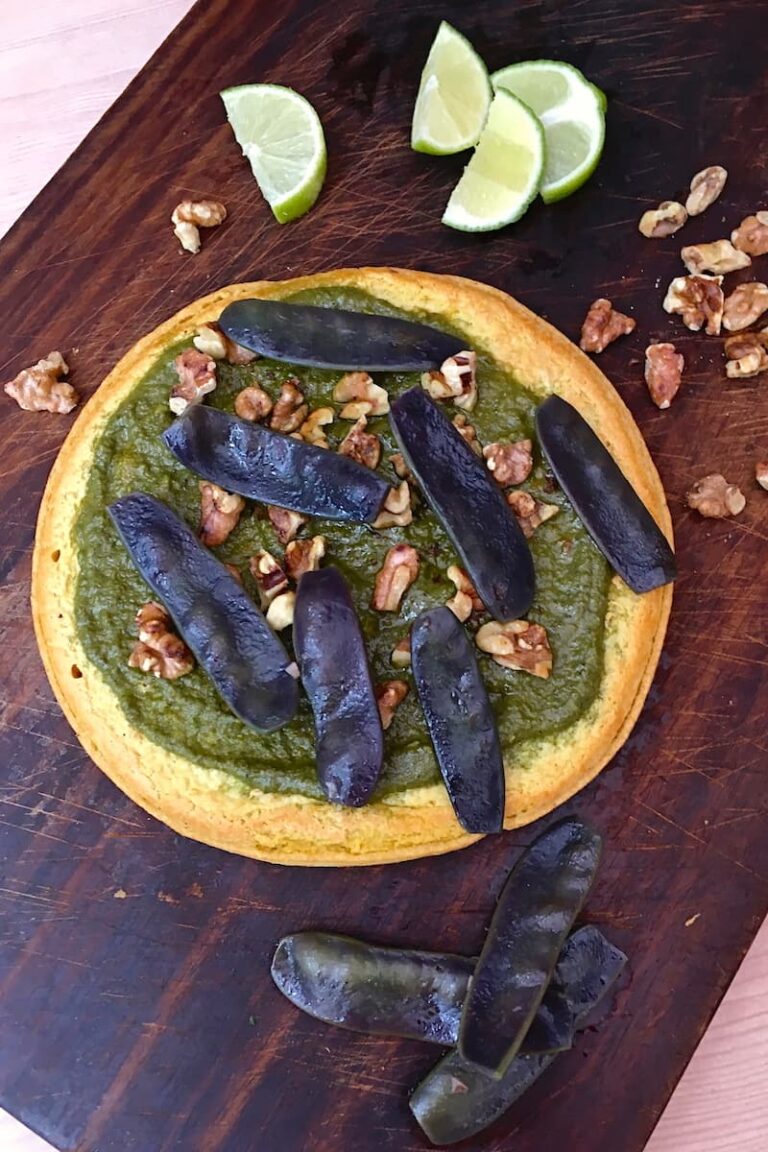 Socca with walnut basil pesto & purple podded peas on a board with walnuts and lime slices.