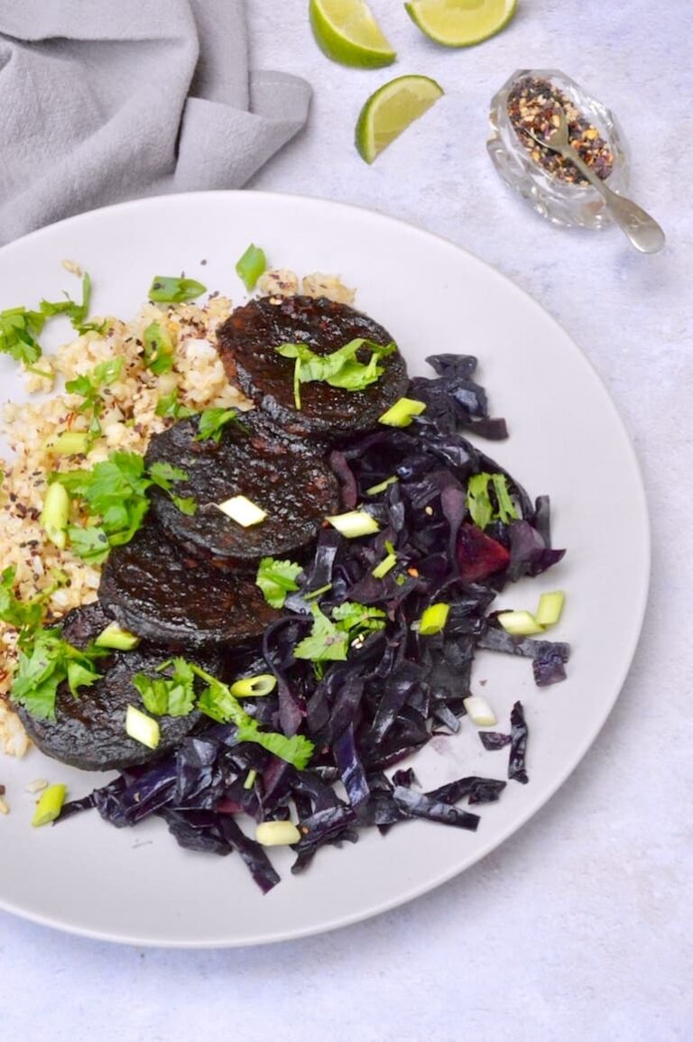 Four slices of sticky gochujang tempeh on a plate with brown rice and red cabbage with coriander and scallions scattered over the top. A grey napkin, lime wedges and a pot of seasoning in the background.
